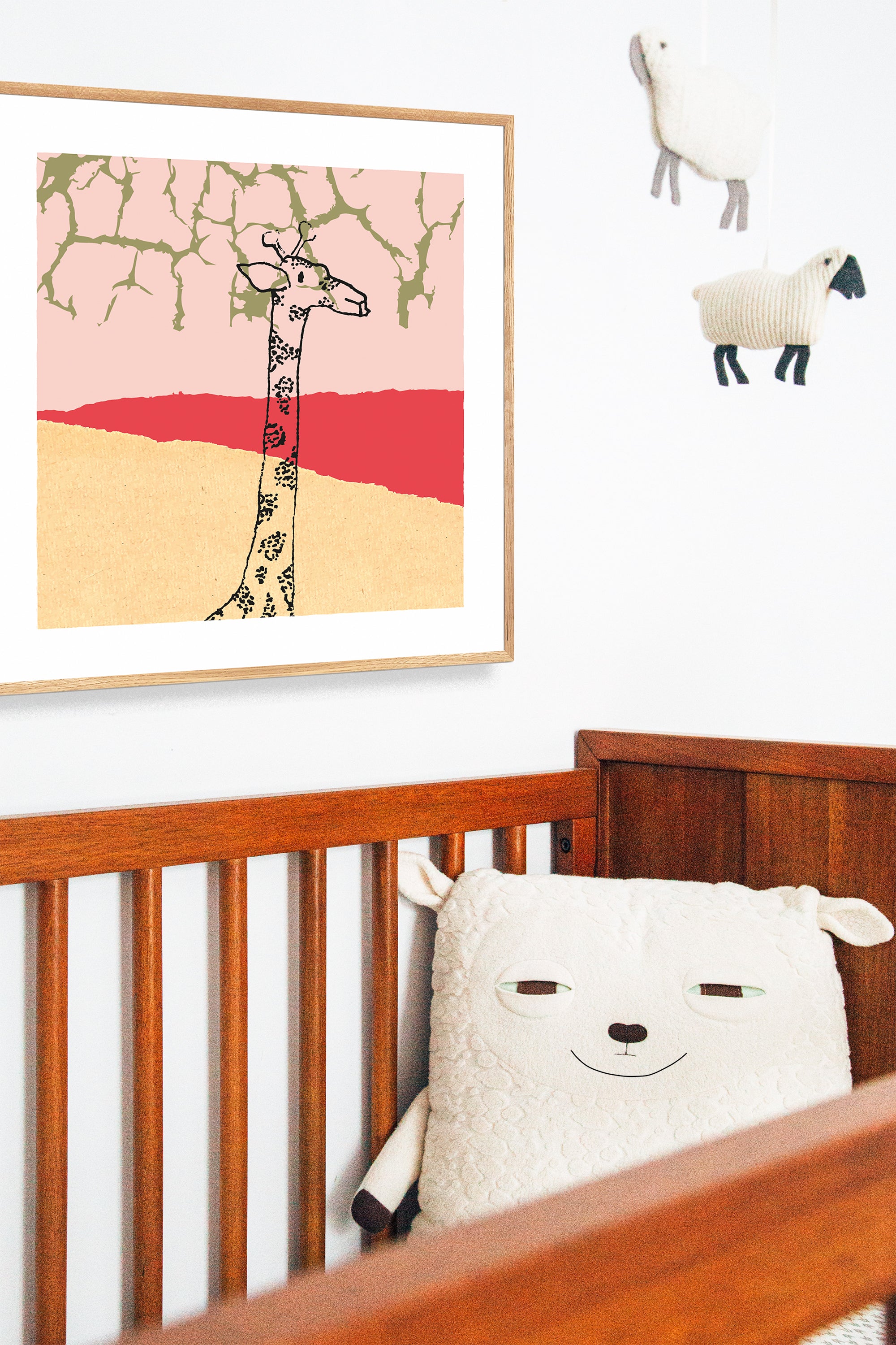 Nursery with cot and sheep mobile and hung art print of giraffe neck and head in black on a pale yellow and red landscape with light pink sky and golden branches reaching from above in a natural wooden frame.